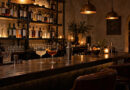 Dunkle Holztheke einer Cocktailbar mit zwei Cocktails, Flaschenregal und warmem Licht. Zum Artikel Bamberg: „Das Schwarze Schaf“ gehört zu den 111 besten Bars Europas